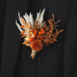 Puede incluir: Un boutonniere de flores secas con acentos blancos, naranjas y marrones. El boutonniere está hecho con hierbas secas, flores y bayas.