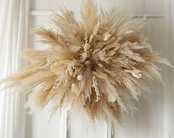 May include: A large, fluffy, dried pampas grass wreath in shades of beige and cream. The wreath is hanging on a white wall.