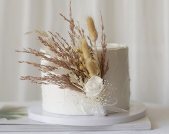 May include: A white cake with a white flower and dried grasses on top. The cake is on a white cake stand and a magazine is underneath.