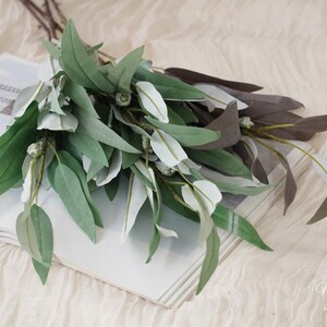 May include: A bouquet of faux eucalyptus leaves in shades of green, white, and brown rests on an open book. The leaves have a realistic appearance, with varying shades and textures. The book is open to a blank page, and the background is a soft, neutral fabric.