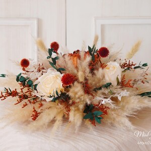 May include: A centerpiece arrangement featuring white roses, red berries, and dried pampas grass. The arrangement is in a rustic, natural style, perfect for a wedding or special event.