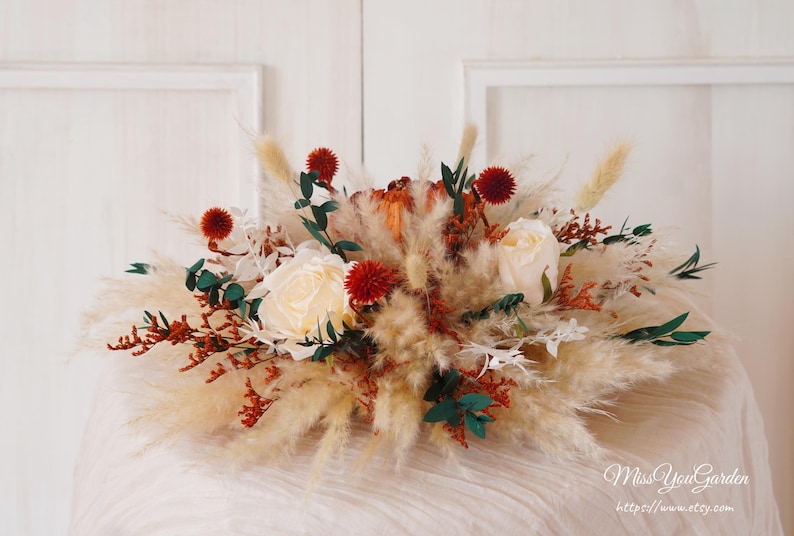 May include: A centerpiece arrangement featuring white roses, red berries, and dried pampas grass. The arrangement is in a rustic, natural style, perfect for a wedding or special event.