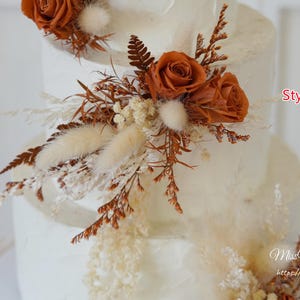 May include: A multi-tiered white cake decorated with rust-colored roses, dried ferns, and cream-colored bunny tail grass. The text Style B is visible in red, and the text MissYouGarden https://www.etsy.com is visible in white script.