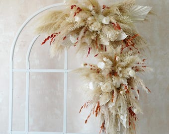 May include: A white metal arch decorated with dried pampas grass, white flowers, and red accents. The arrangement is full and lush, creating a natural and elegant aesthetic. Perfect for weddings or home decor.