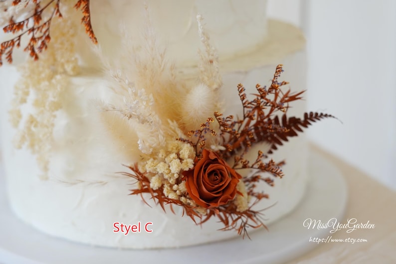 May include: A white tiered cake decorated with dried floral arrangements featuring a rust-colored rose, beige pampas grass, cream-colored baby's breath, and brown fern fronds. The text "Styel C" and "MissYouGarden https://www.etsy.com" are visible.