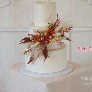 May include: A three-tiered white cake with a textured finish, decorated with a cluster of burnt orange roses, dried ferns, and pampas grass. The text Styel D and MissYouGarden https://www.etsy.com are visible on the right side of the image.
