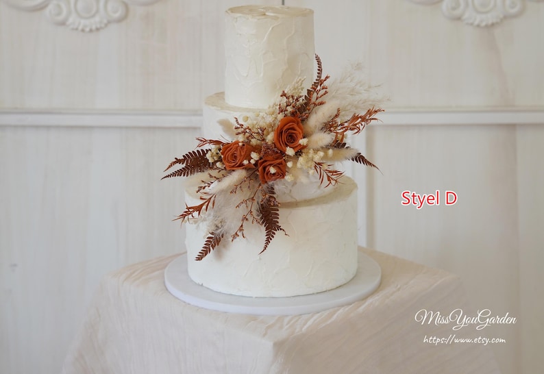 May include: A three-tiered white cake with a textured finish, decorated with a cluster of burnt orange roses, dried ferns, and pampas grass. The text Styel D and MissYouGarden https://www.etsy.com are visible on the right side of the image.