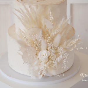 May include: A two-tiered white wedding cake decorated with dried pampas grass, white flowers, and other dried botanicals. The cake is on a white cake stand. The text "Miss You Garden" and "https://www.etsy.com" is visible at the bottom of the image.
