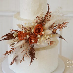May include: A three-tiered white cake decorated with a cluster of dried flowers including burnt orange roses, cream-colored pampas grass, and brown fern fronds. The cake has a textured white frosting finish and is adorned with the text MissYouGarden.