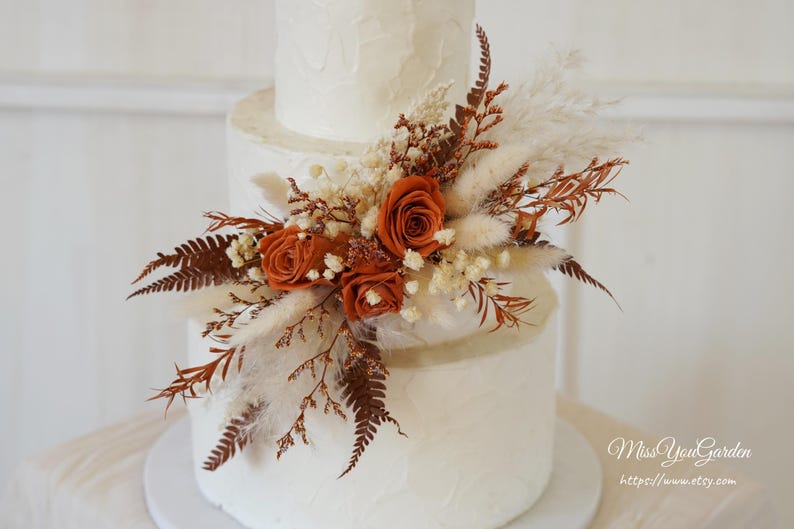 May include: A three-tiered white cake decorated with a cluster of dried flowers including burnt orange roses, cream-colored pampas grass, and brown fern fronds. The cake has a textured white frosting finish and is adorned with the text MissYouGarden.