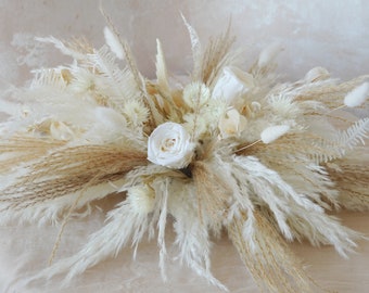May include: A white dried flower arrangement with pampas grass, white roses, and other dried flowers.