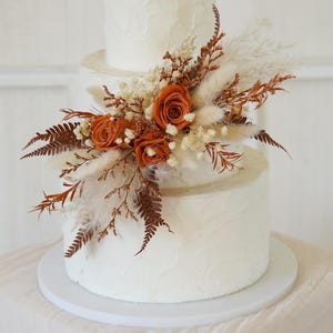 May include: A three-tiered white cake with textured frosting, decorated with a cluster of dried rust-colored roses, white pampas grass, small white filler flowers, and brown fern fronds. The text Miss You Garden https://www.etsy.com is visible.