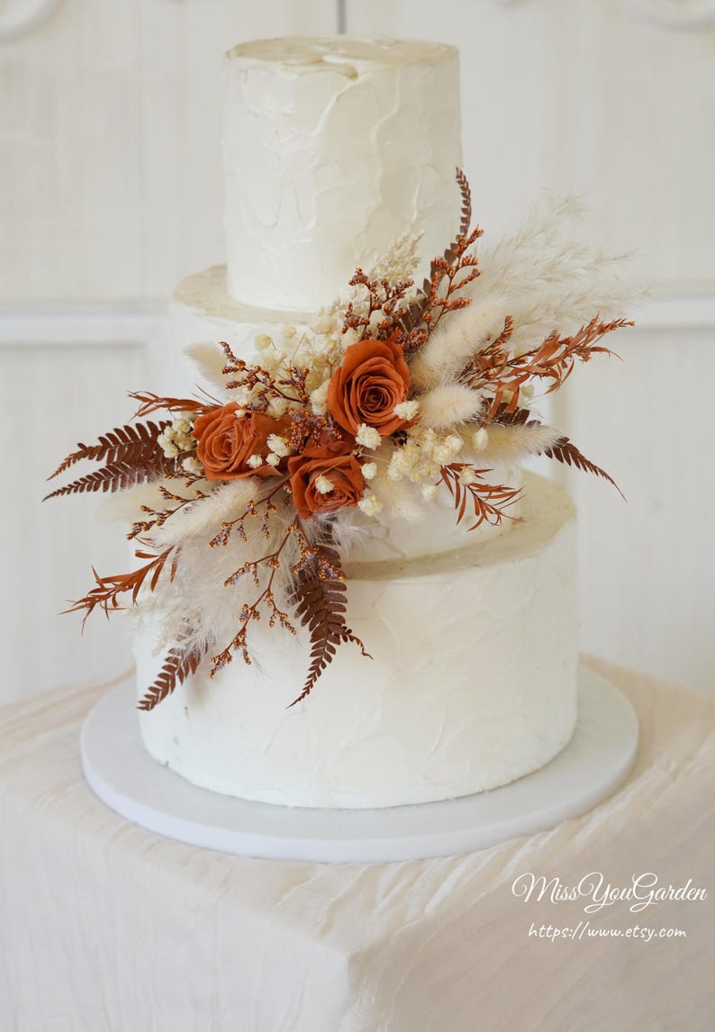 May include: A three-tiered white cake with textured frosting, decorated with a cluster of dried rust-colored roses, white pampas grass, small white filler flowers, and brown fern fronds. The text Miss You Garden https://www.etsy.com is visible.