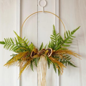 Peut inclure: Une couronne décorative avec un anneau en métal doré, des feuilles de fougère vertes, des tranches d'orange séchées et des herbes de couleur beige. Un ruban transparent est suspendu en bas. La couronne est affichée sur un fond blanc.