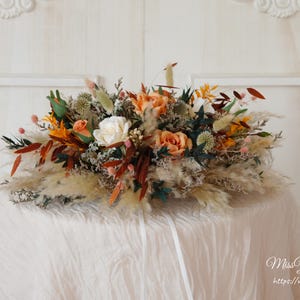 Könnte beinhalten: Ein Blumenarrangement aus getrockneten Blumen mit pfirsichfarbenen Rosen, weißen Blüten und orangefarbenem, grünem und braunem Laub. Das Bouquet steht auf einer weißen, drapierten Oberfläche. Enthält Pampasgras.