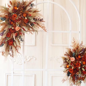 Könnte beinhalten: Arrangement aus getrockneten Blumen und Pampasgras in Orange, Rot und Beige. Das Blumenarrangement befindet sich vor einem weißen Hintergrund mit einem dekorativen Bogen und einer weißen Tür.
