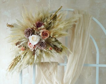 May include: A dried floral arrangement with pampas grass, dried palm leaves, and blush pink, mauve, and white roses. The arrangement is mounted on a white metal archway with a sheer white fabric draped over it.