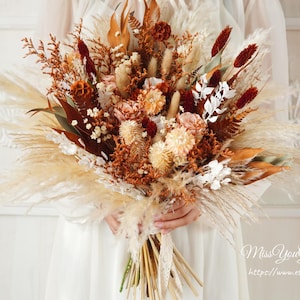 Può includere: Un bouquet di fiori secchi con erba della pampa, eucalipto essiccato e altri fiori secchi in tonalità di marrone, beige e rosso. Il bouquet è legato con un nastro di pizzo bianco.