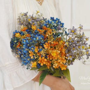 May include: A bouquet of artificial flowers in shades of blue, orange, and purple. The flowers are held in a person's hands. The bouquet has green leaves. The background is white.