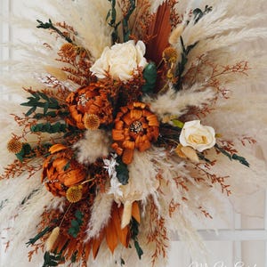 May include: A dried floral arrangement with a mix of pampas grass, dried roses, and other dried flowers in shades of brown, cream, and white. The arrangement is in a rustic style and would be a beautiful addition to any home decor.