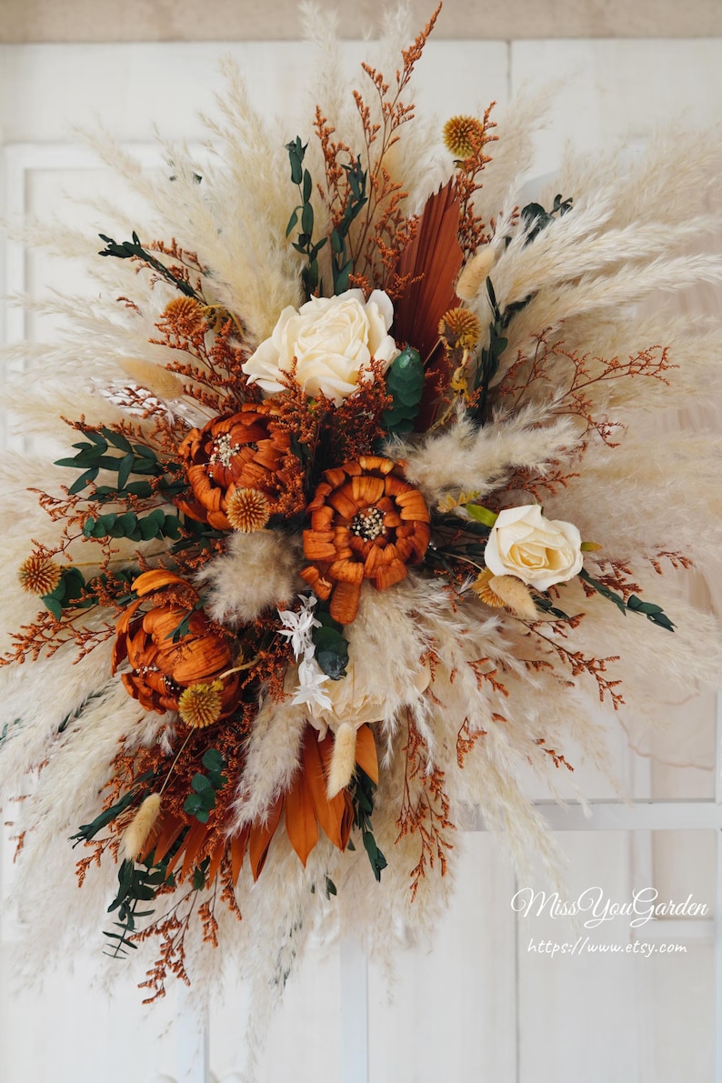 May include: A dried floral arrangement with a mix of pampas grass, dried roses, and other dried flowers in shades of brown, cream, and white. The arrangement is in a rustic style and would be a beautiful addition to any home decor.