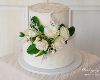 Op de afbeelding: Een witte taart met twee lagen en getextureerde frosting, versierd met witte rozen, groene bladeren en delicate takjes. De taart staat op een witte taartstandaard, perfect voor een bruiloft of viering.