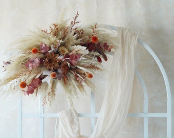 May include: A dried floral arrangement featuring pampas grass, orange globe thistle, and burgundy eucalyptus. The arrangement is displayed on a white metal arch with a cream-colored fabric draped over it. The background is a textured, off-white wall.
