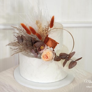 May include: A two-tiered white cake decorated with dried flowers, pampas grass, and a gold ring. The cake features orange, brown, and peach-colored floral accents. The cake sits on a white cake stand.