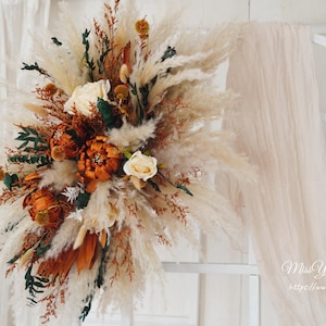 May include: A dried floral arrangement with pampas grass, dried roses, and other dried flowers in shades of brown, white, and green. The arrangement is displayed against a white background.
