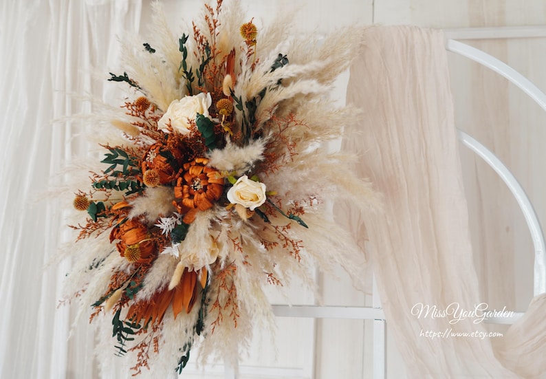 May include: A dried floral arrangement with pampas grass, dried roses, and other dried flowers in shades of brown, white, and green. The arrangement is displayed against a white background.