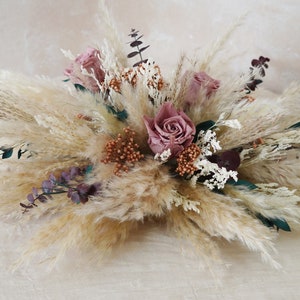May include: A dried flower arrangement featuring a mix of pampas grass, dried roses, and eucalyptus. The roses are a soft pink colour, and the pampas grass is a light beige colour. The arrangement is perfect for adding a touch of natural beauty to any room.