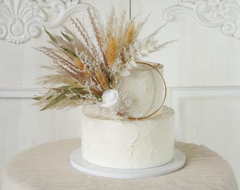 May include: A two-tiered white wedding cake decorated with dried pampas grass and a gold ring. The cake is on a white cake stand and a beige tablecloth.