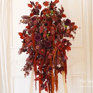 Könnte beinhalten: Ein großer Blumenstrauß mit tiefroten und burgunderfarbenen Blumen und Blättern. Das Arrangement umfasst Rosen, Beeren und kaskadenartige Elemente, die eine reichhaltige, strukturierte Darstellung schaffen. Der Hintergrund ist eine weiße Wand und ein Vorhang.
