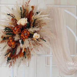 May include: A dried floral arrangement with pampas grass, dried flowers, and greenery. The arrangement is in shades of brown, white, and green. It is displayed on a white background.