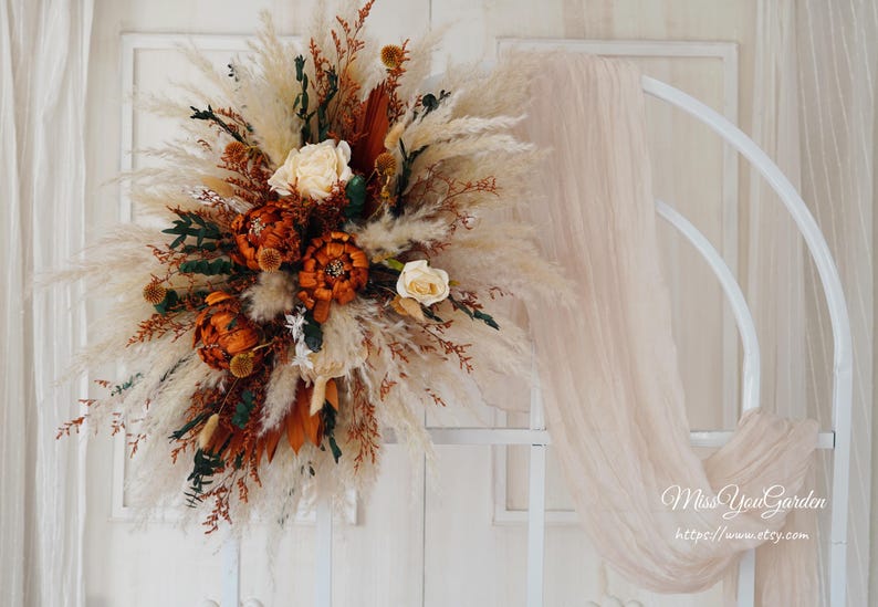May include: A dried floral arrangement with pampas grass, dried flowers, and greenery. The arrangement is in shades of brown, white, and green. It is displayed on a white background.