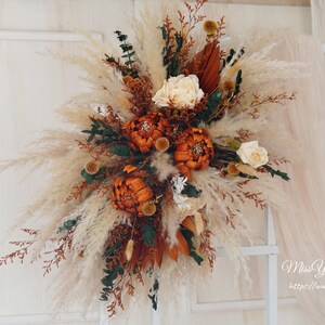May include: A dried floral arrangement with pampas grass, dried flowers, and greenery in shades of brown, orange, and white. The arrangement is hanging on a white wall.