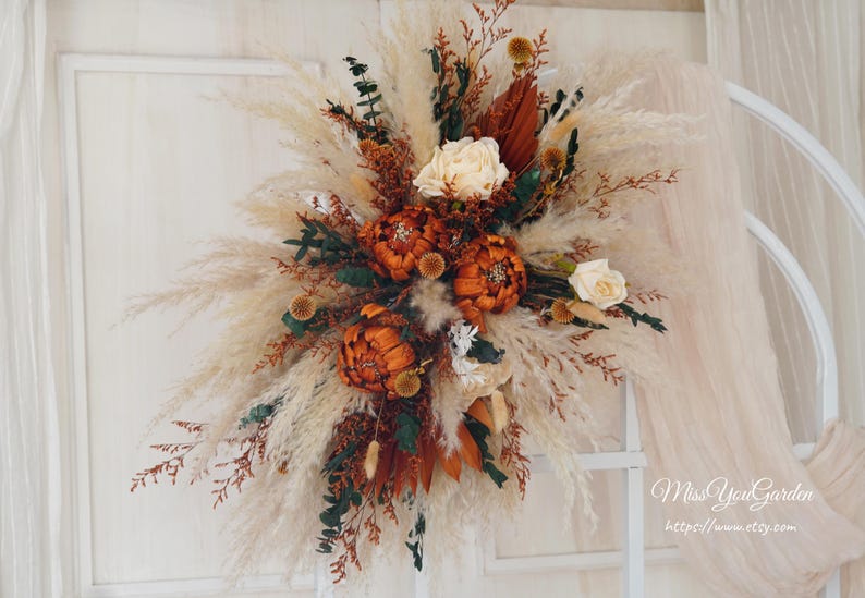 May include: A dried floral arrangement with pampas grass, dried flowers, and greenery in shades of brown, orange, and white. The arrangement is hanging on a white wall.
