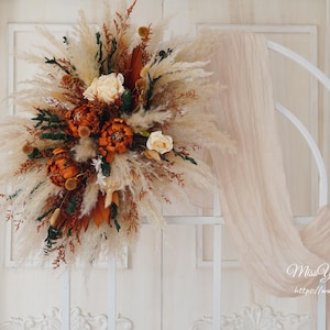 May include: A white metal archway decorated with a large bouquet of dried pampas grass, dried flowers, and dried foliage in shades of brown, orange, and cream. The archway is decorated with a sheer white fabric.