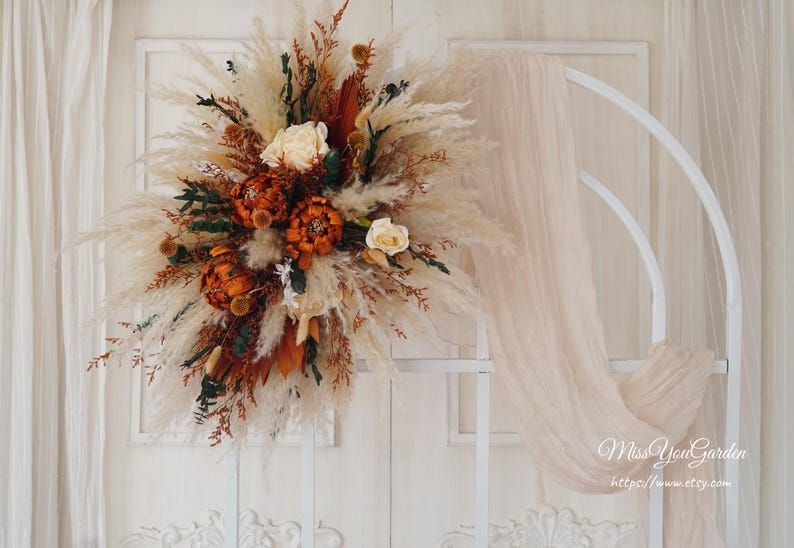May include: A white metal archway decorated with a large bouquet of dried pampas grass, dried flowers, and dried foliage in shades of brown, orange, and cream. The archway is decorated with a sheer white fabric.
