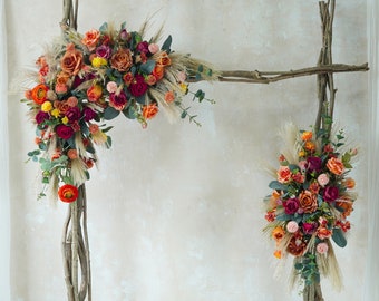 May include: A rustic wooden archway decorated with dried grasses and a mix of orange, red, and pink flowers. The archway is perfect for a wedding or other special event.