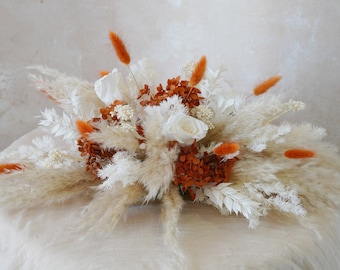 May include: A dried floral arrangement featuring white pampas grass, white roses, and orange dried hydrangeas.