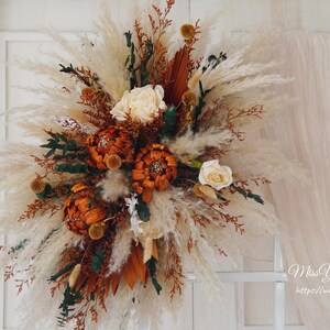 May include: A dried floral arrangement with white pampas grass, dried orange flowers, and brown dried leaves. The arrangement is in a white vase and is on a white background.