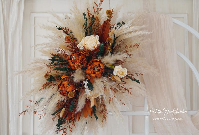 May include: A dried floral arrangement with white pampas grass, dried orange flowers, and brown dried leaves. The arrangement is in a white vase and is on a white background.