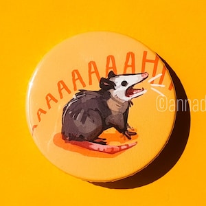 Peut inclure: Un bouton jaune avec un opossum de dessin animé criant "AAAAAH!" sur un fond blanc. L'opossum est brun et gris avec un visage blanc et un nez rose.