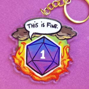 Può includere: Un portachiavi con un dado D20 che mostra il numero 1, circondato da fiamme. Sopra il dado, una nuvoletta dice "This is Fine." Il design include una catena e un anello dorati, su uno sfondo viola.