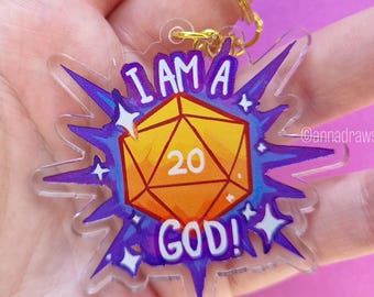 Könnte beinhalten: Ein klarer Acryl-Schlüsselanhänger mit einem goldfarbenen Ring. Der Schlüsselanhänger zeigt ein gelb-oranges zwanzigseitiges Würfeldesign mit der Zahl 20 in der Mitte. Der Text "I AM A GOD!" ist in Lila um den Würfel geschrieben.