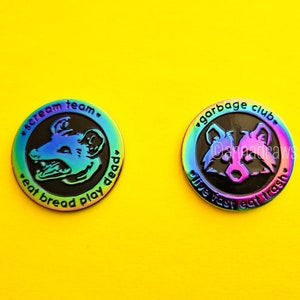 May include: Two iridescent enamel pins with black backgrounds. The first pin features a cartoon opossum with the text "scream team. eat bread play dead". The second pin features a cartoon raccoon with the text "garbage club. live fast eat trash".