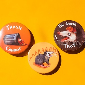 Könnte beinhalten: Drei runde Knöpfe mit bunten Designs. Der erste Knopf ist orange und zeigt einen schwarzen Mülleimer mit herausfallenden Blumen. Der Text auf dem Knopf lautet "Trash Cannot". Der zweite Knopf ist gelb und zeigt ein schwarz-weißes Opossum mit dem Text "AAAAAH". Der dritte Knopf ist schwarz und zeigt ein weißes Opossum mit roten Blumen im Haar. Der Text auf dem Knopf lautet "Be Gone Thot!"
