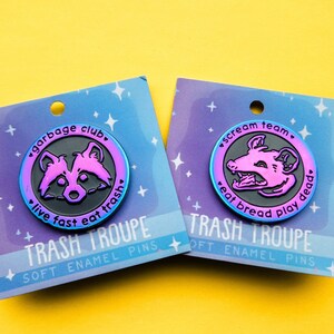 Op de afbeelding: Twee emaillepins met een zwarte en iriserende afwerking. De eerste pin heeft een wasbeer met de tekst "garbage club. live fast eat trash". De tweede pin heeft een opossum met de tekst "scream team. eat bread play dead". Beide pins hebben de tekst "TRASH TROUPE SOFT ENAMEL PINS".