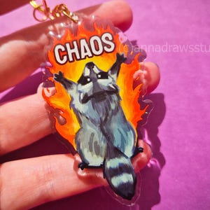May include: A clear acrylic keychain featuring a cartoon raccoon with its arms raised, surrounded by orange and yellow flames. The word CHAOS is printed in white capital letters above the raccoon. The keychain includes a gold-colored metal ring.